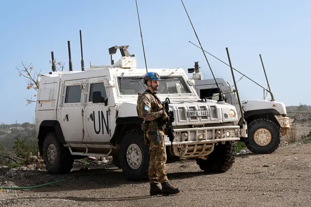 أحد أفرد قوة الأمم المتحدة الموقتة العاملة في جنوب لبنان (يونيفيل) بلباس عسكري وخوذة زرقاء، يقف إلى جانب مركبة بيضاء عليها حروف دالة على الأمم المتحدة. الصورة التقطت نهاراً.