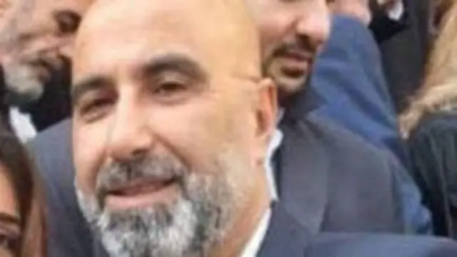 بيار معوض يظهر من الأعلى يبدو وكأنه يرتدي بدلة رسمية رمادية اللون، بشرته قمحية اللون ولديه لحية رمادية اللون وليس لديه شعر وحوله عدد من الأشخاص.
