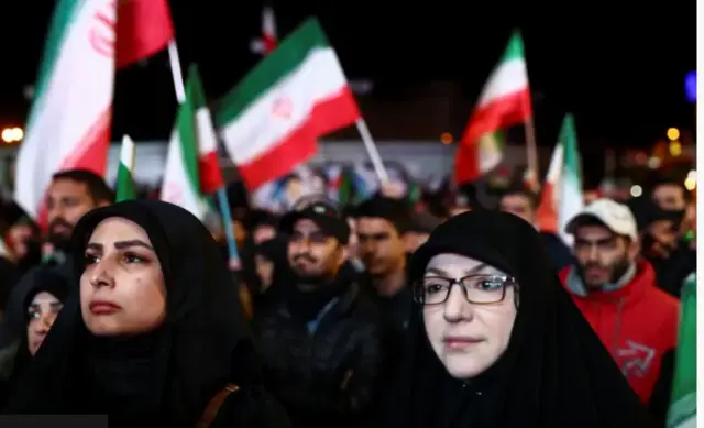 Ifoto igaragaza amasura y'abantu benshi, barimo n'abagore, bari mu muhanda, bamwe bafite amabendera.