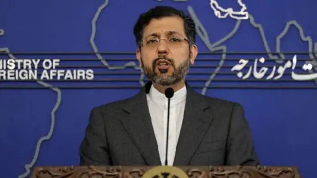 Umugabo urimo kuvugira mu ndangururamajwi, yambaye ikositimu y'umukara n'ishati y'umweru.