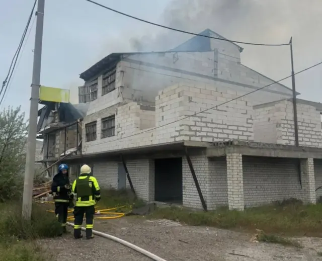 Пожарные около кирпичного дома в три этажа, с последнего этажа валит дым.