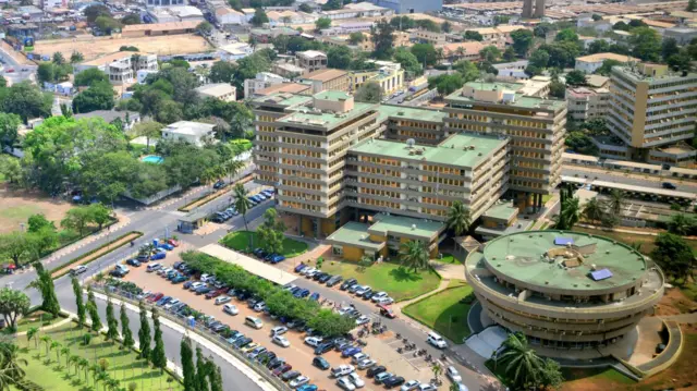Ifoto ifatiwe hejuru igaragaza inyubako nyinshi ndende n'izindi nzu, hamwe n'imodoka nyinshi ziparitse hanze.