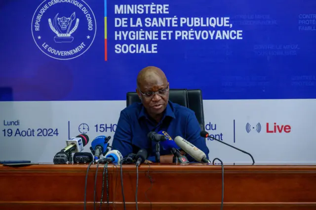 Minisitiri w’ubuzima wa DRC Dr Samuel-Roger Kamba mu kiganiro n’abanyamakuru i Kinshasa, ku itariki ya 19 Kanama (8) mu mwaka wa 2024. Yambaye ishati y'ubururu y'amaboko maremare, imbere ye hari indangururamajwi nyinshi.