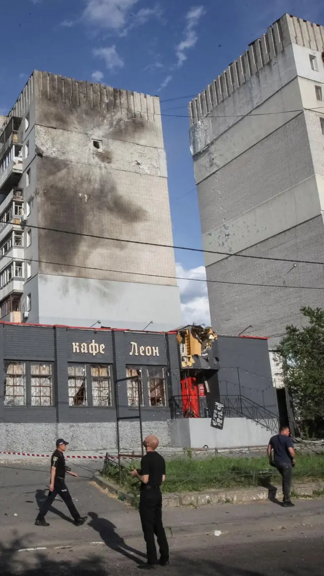 Люди в черной форме ходят вокруг серых домов, в одном из них пробоина в боковой стене, в нижнем этаже - кафе "Леон"