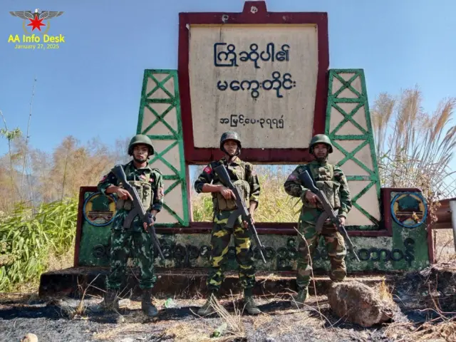 ကြိုဆိုပါ၏ မကွေးတိုင်း အမြင့်ပေ ၃၇၇၅ လို့ရေးထားတဲ့ မြို့အဝင်ကျောက်တိုင်ရှေ့မှာ သေနတ်ကိုင်ရပ်နေတဲ့ အေအေတပ်ဖွဲ့ဝင် ၃ ဦး
