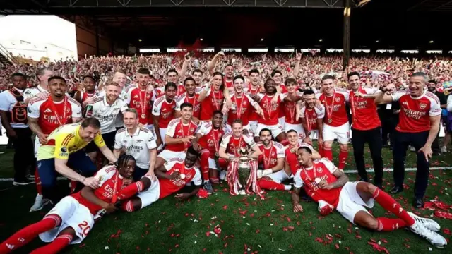 Abakinnyi ba Arsenal, bamwe baryamye mu byatsi, abandi basutamye, naho abandi bahagaze, bakikije igikombe kiri hasi. Bambaye imyenda y'umutuku n'umweru, inyuma yabo hari abantu benshi bahagaze.