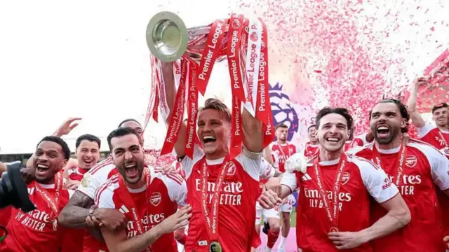 Umukinnyi wa Arsenal azamuye igikombe kiriho udutambaro tw'umutuku n'umweru twanditseho "Premier League". Ari kumwe n'abandi bakinnyi benshi basamye bishimye, bambaye imyenda y'umutuku n'umweru. Inyuma yabo hari ibishashi bitukura by'imiriro yo mu birori.