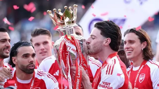 Umukinnyi wa Arsenal arimo gusoma igikombe cya Premier League, akikijwe n'abandi bakinnyi.