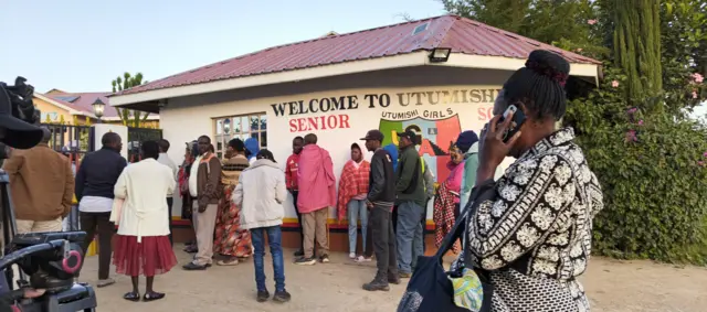 Itsinda ry'abantu benshi bahagaze imbere y'inyubako y'ishuri muri Kenya. Harimo abagabo n'abagore, umugore umwe aboneka arimo kuvugira kuri telefone.
