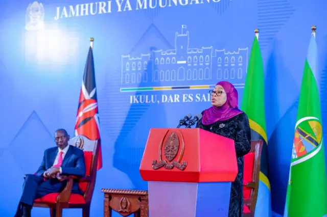Perezida wa Kenya William Ruto yicaye arangamiye Perezida wa Tanzania Samia Suluhu ahagaze arimo kuvuga ijambo. Inyuma ya Ruto hari ibendera rya Kenya, mu gihe inyuma ya Samia hari ibendera rya Tanzania.