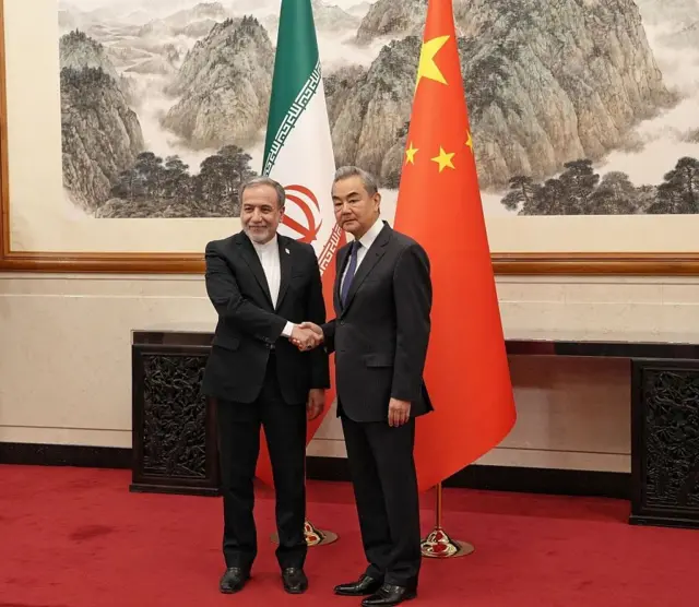 Minisitiri w'ububanyi n'amahanga wa Iran Abbas Araghchi (ibumoso) na Minisitiri w'ububanyi n'amahanga w'Ubushinwa Wang Yi, i Beijing, ku itariki ya 6 Gicurasi (5) mu 2026. Bahagaze ku mucyeka utukura bahanye ikiganza, bambaye amakositimu y'umukara, inyuma hari amabendera y'ibihugu byombi.