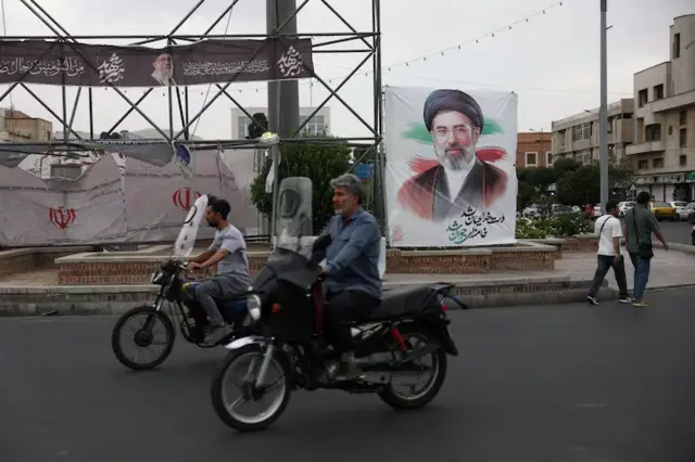 Abantu bariko bagendesha amapikipiki baca iruhande y’ifoto nini y’Umuyobozi w’ikirenga, Mojtaba Khamenei, mw’ibarabara ry’i Tehran, muri Iran, ku wa 7 Ruheshi (6) 2026.