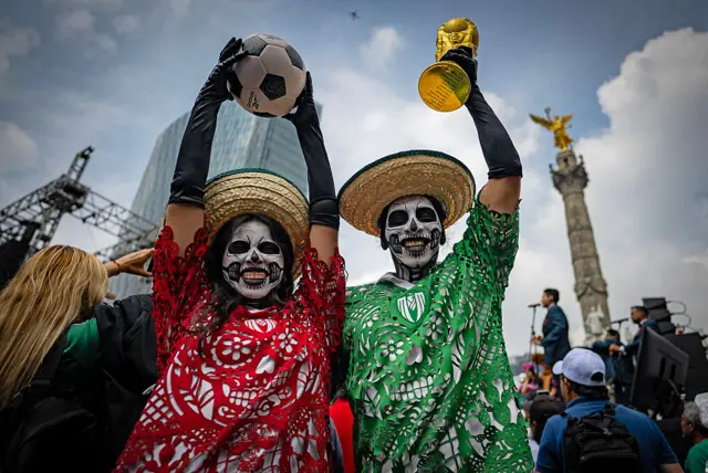 Dos personas con el rostro pintado, con camisetas coloridas en verde y rojo, alzan la Copa del Mundo y un balón frente al Monumento a la Independencia en Ciudad de México