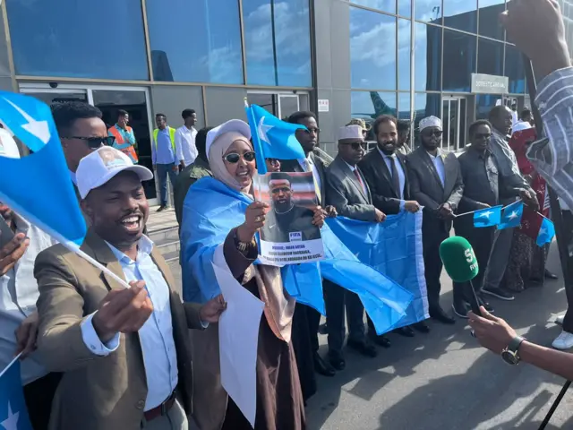 Imbaga y'abantu bafite amabendera ya Somalia y'ubururu bwerurutse, batonze umurongo imbere y'inyubako y'ibirahure, bamwe bamwenyura. Imbere yaho hari ukuboko kw'umuntu ufashe indangururamajwi.