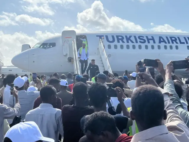 Imbaga y'abantu bareba Omar Artan amanuka ku madarage y'umuryango w'indege ya kompanyi yo muri Turukiya. Barimo abari gufotoresha 'cameras' z'abanyamakuru, n'abafotoresha telefone, bateye umugongo muri 'camera'.