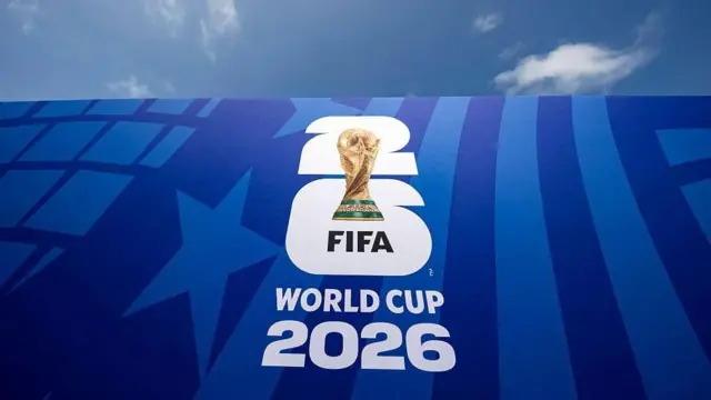 Ifoto y’ikimenyetso c’igikombe c’isi ca FIFA 2026, yanditsweko mu ndome zera. Irimwo igikombe c’umupira w’amaguru c’inzahabu gifise amabara y’icatsi kibisi ku ruhande rwo hepfo.
