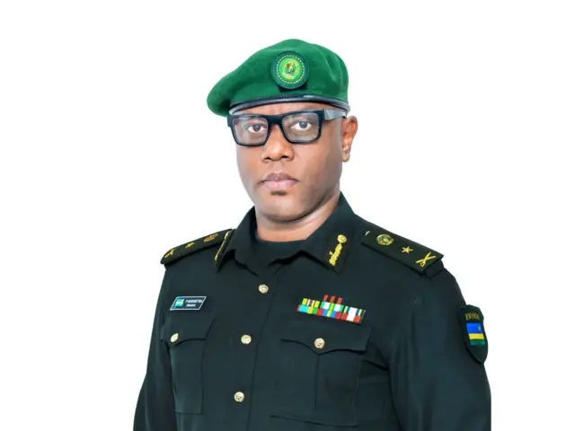 Brig Gen Patrick Karuretwa yambaye ishati ya gisirikare y'ibara ry'icyatsi kibisi gitsitse, iriho amapeti. Imbere yambayemo umupira w'icyatsi kibisi. Yambaye n'ingofero y'icyatsi kibisi n'indorerwamo.