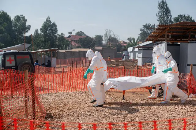 Abakora mu buvuzi batwaye umurambo w'umuntu wishwe na Ebola bawujyanye mu buruhukiro bw'ikigo nderabuzima cya Nyankunde, i Bunia, mu ntara ya Ituri, ku wa gatandatu ushize, kugira ngo benewabo bawusezereho bwa nyuma. Bambaye imyenda yabugenewe y'umweru ibapfutsemo umubiri wose, ndetse n'ingofero z'umweru, banambaye udupfukantoki hamwe n'inkweto. Barimo kugenda mu mbuga y'ivuriro irimo amabuye, ikikijwe n'inzitiro z'ibara rya 'orange'. Ifoto yo ku itariki ya 13 Kamena (6) mu mwaka wa 2026.