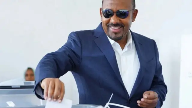 Abiy Ahmed yambaye ikositime y'ubururu, ishati yera, n'amarori y’izuba, ariko aramwenyura mu gihe ariko ashira ikarata y’itora mw’isandugu