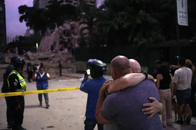 Dos hombres se abrazan mientras en el fondo hay daños por el terremoto