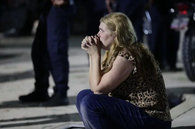 Una mujer sentada sobre una acera en la noche. Tiene las manos junto a su boca y una expresión de angustia.