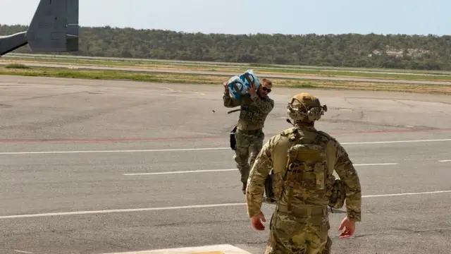 Un oficial estadounidense carga un paquete sobre su hombro por la pista de aterrizaje del aeropuerto. Se ve de espaldas a otro agente de EE.UU.