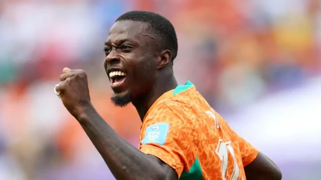 Nicolas Pépé wambaye umwambaro w'ibara rya orange agaragara ari mu kibuga yishimye cyane ari guseka yazamuye akaboko kamwe yafunze igipfunsi