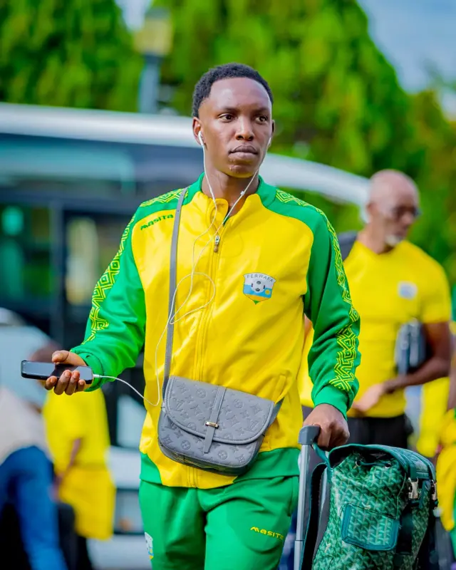 Umukinnyi w'ikipe y'igihugu y’umupira w’amaguru y'u Rwanda, Amavubi, arimo gutambuka, afite telefone mu kiganza, n'udukoresho two kumvisha twa telefone. Afashe aho gufatira h'ivalisi iriho igikapu gito hejuru na cyo agifashe umushumi, ndetse afite ikindi gikapu gito ahetse kiri mu nda. Yambaye imyenda y'icyatsi kibisi n'umuhondo y'ijaketi n'ikabutura, iriho ikirango cya FERWAFA. Inyuma ye hari imodoka itagaragara neza hamwe n'abandi bantu bataboneka neza.