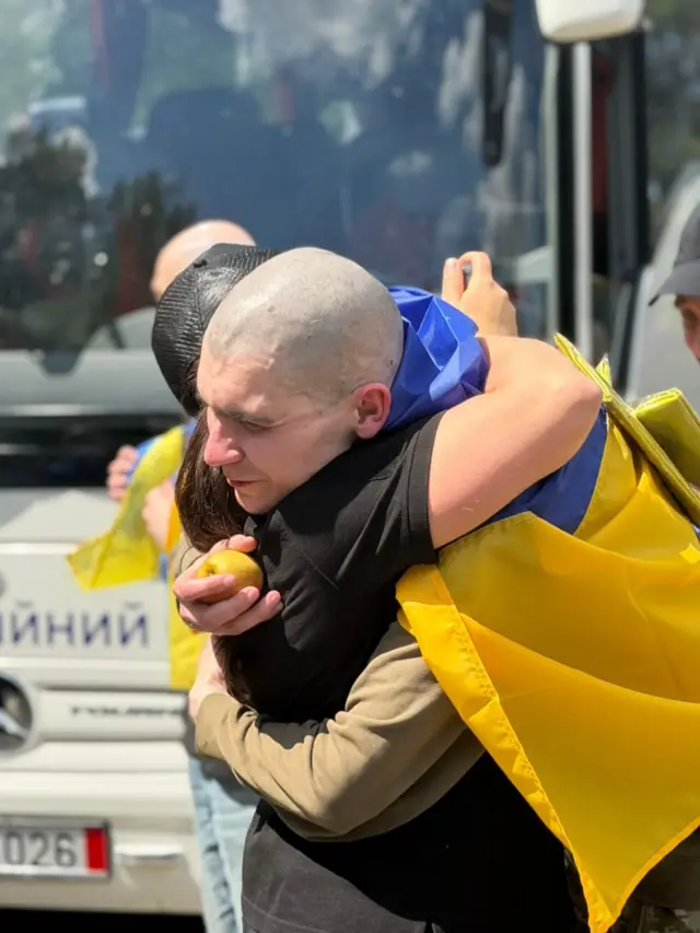 Мужчина, закутанный в украинский флаг, обнимает женщину.