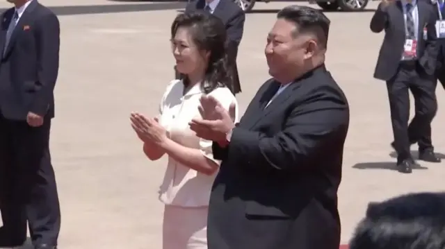 Kim Jong Un n'umugore we Ri Sol Ju bakoma amashyi ndetse bamwenyura. Barareba umuntu utaboneka mu ifoto. Inyuma n'iruhande rwabo hari abashinzwe umutekano bambaye amakositimu.