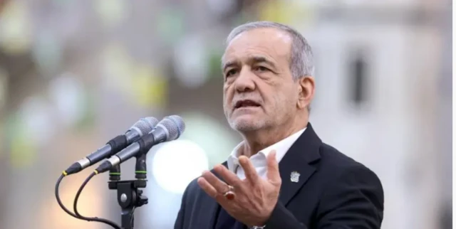 الرئيس الإيراني مسعود بزشكيان، يرفع يده أثناء إلقاء خطاب.. وهو يرتدي بدلة داكنة اللون ويتحدث عبر ميكروفونين.