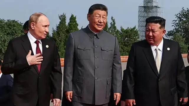 Uvuye ibumoso ujya iburyo: Perezida w'Uburusiya Vladimir Putin, Perezida w'Ubushinwa Xi Jinping n'umutegetsi wa Koreya ya Ruguru Kim Jong Un, barahagararanye, bambaye amakositimu yijimye. Putin na Kim barareba ku mpande, mu gihe Xi areba imbere.