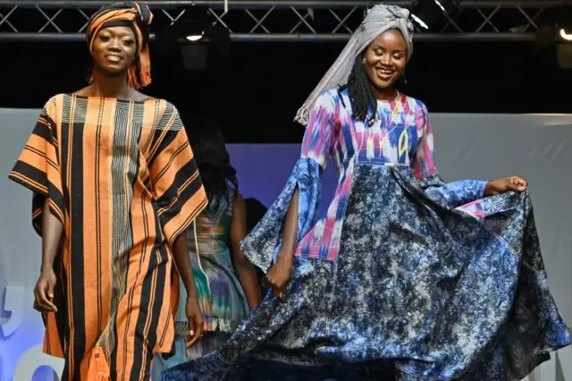 Abagore babiri bambaye imyenda y'amakanzu idoze mu bitenge barimo gutambuka imbere ahantu higiye hejuru, barimo kumwenyura. Inyuma haraboneka undi mugore wambaye umwenda wo mu gitenge, atambuka agana mu kindi cyerekezo.
