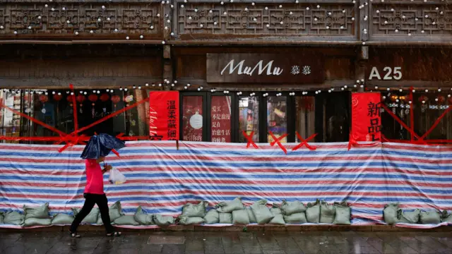 浙江溫州街頭一商店以帆布和沙包圍封正門，一位身穿鮮紅色上衣的男子打著雨傘，拿著購物走過（11/7/2026）