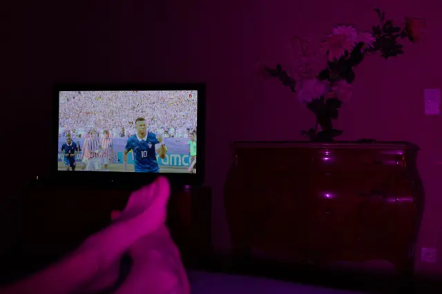 Televisor exibindo jogo da Copa com Mbappé na tela, e pés de espectador no primeiro plano