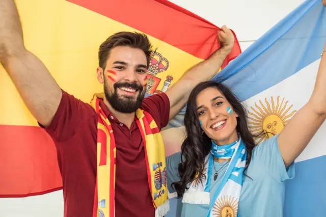 Un groupe de jeunes supporters de football de différentes origines ethniques acclament leur équipe lors de la Coupe du monde. Ils brandissent des drapeaux de leurs équipes nationales et ont le visage peint de couleurs vives.