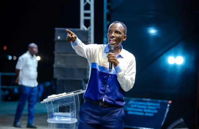 Chris Ndikumana mw'ishati ifise ibara ryera, iryirabura n'ubururu, agoroye ukuboko kw’iburyo nk’uwuriko yerekana ikintu, mu kundi afise 'micro', ariko aravuga(ifoto mbarankuru yo mu bubiko)