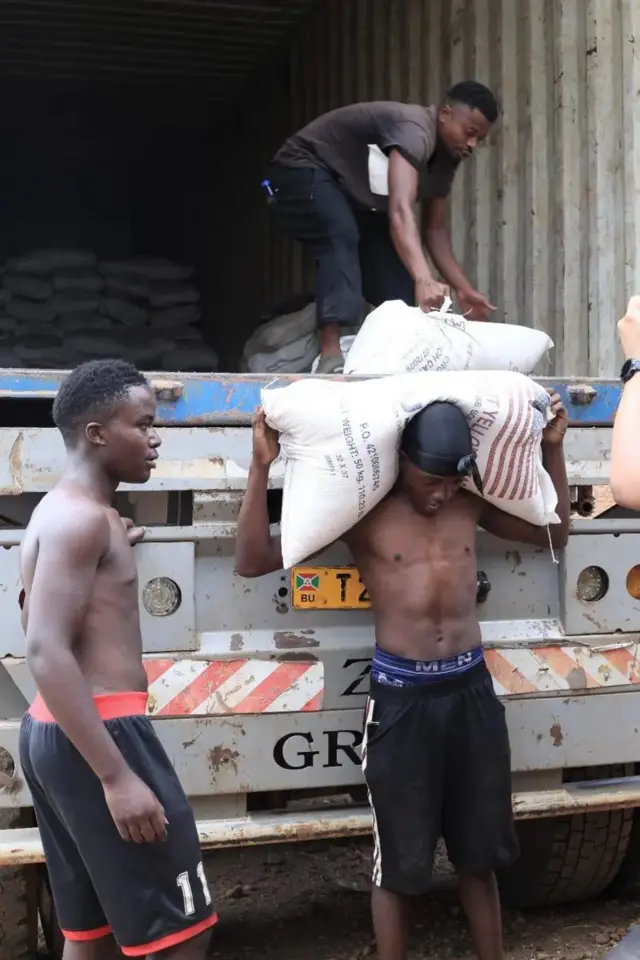 Abagabo batatu barimo gupakurura imifuka mu ikamyo. Babiri bari hasi batambaye mu gatuza, mu gihe imbere mu modoka inyuma hahagazemo undi mugabo