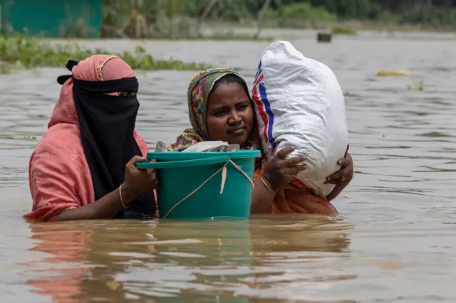 বুক সমান বন্যার পানিতে দুই জন নারী। একজনের কাঁধে একটি বস্তা, আরেকজনের হাতে একটি সবুজ প্লাস্টিকের বালতি