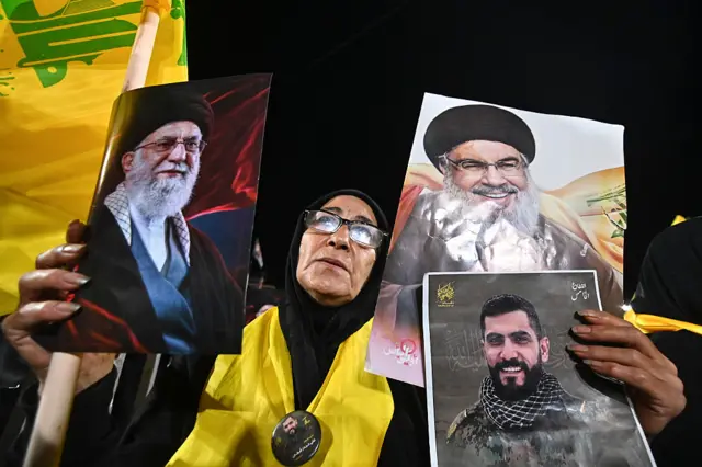 امرأة باللباس الأسود والحجاب تحمل صورة لخامنئي وصورة لحسن نصرالله وصورة لمقاتل في حزب الله وخلفها علم لحزب الله