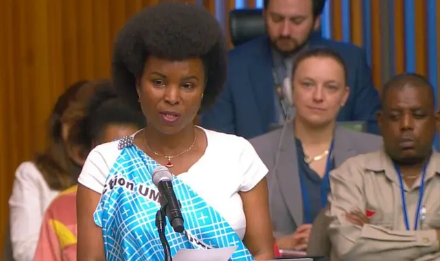 Angeline Ndayishimiye ariko aravuga, yambaye imvutano n'agapira kera, inyuma yiwe haboneka abandi bantu