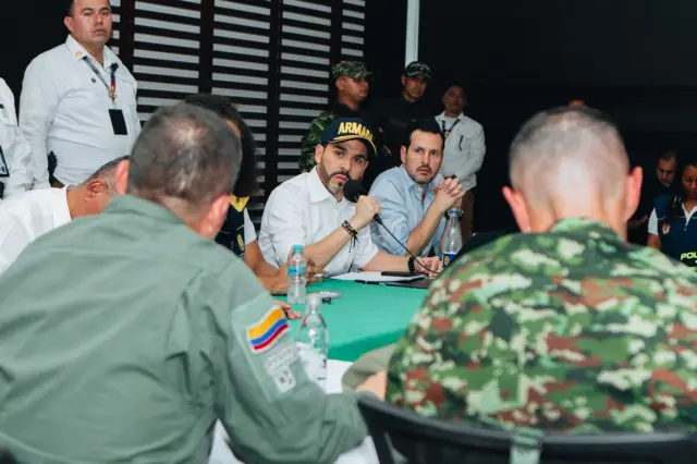 Hombres sentados alrededor de una mesa, algunos de espaldas, otros de frente, vestidos muchos de militares. De la Espriella está en el centro de la imagen con gorra, camisa remangada y micrófono en la mano.
