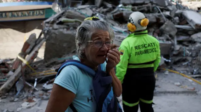 una mujer mayor en primer plano llora mientras se lleva las manos a la cara. Tiene un rescatista detrás