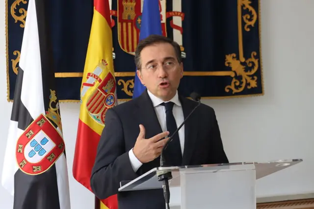 El ministro de Asuntos Exteriores, Unión Europea y Cooperación, José Manuel Albares, habla con los medios de comunicación vestdo de traje de chaqueta y corbata, de pie, tras un atril con micrófono. Detrás hay una bandera de España y otra de Ceuta. En la pared, un escudo de España.