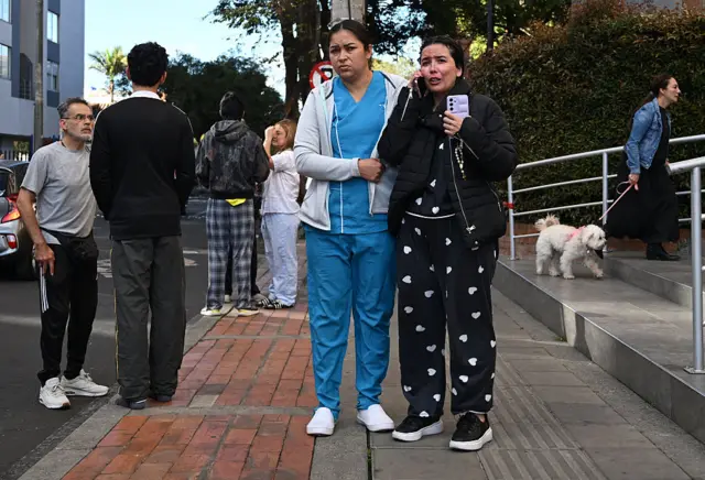 Una mujer vestida con un pijama negro habla por teléfono en la calle con cara de preocupacón, mientras una mujer que parece tener uniforme de enfermera está parada a su lado.