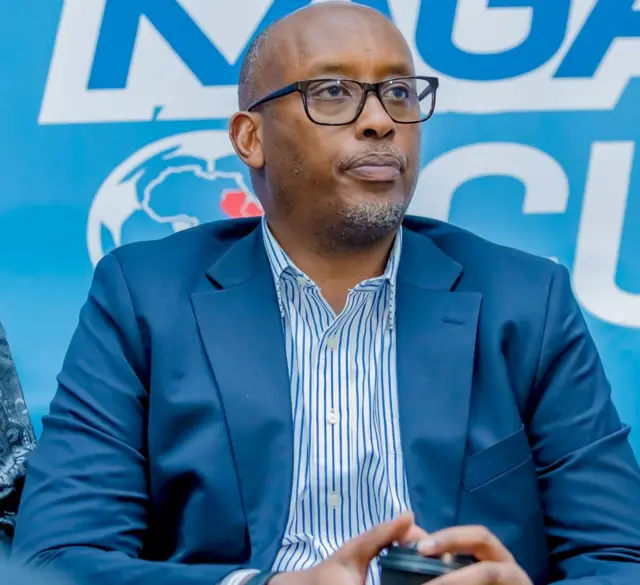 Umuyobozi wa FERWAFA Shema Fabrice yambaye ikositimu y'ubururu n'indorerwamo areba imbere ahagana hejuru. Imbere y'ikoti yambayemo ishati y'umweru n'ubururu. Inyuma ye hari icyapa cy'ubururu cyanditseho mu mweru n'ubururu.