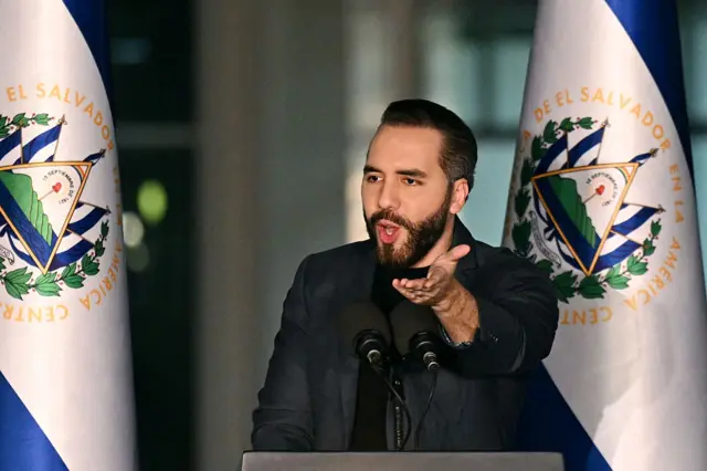 Nayib Bukele, vestido con un traje negro, está de pie ante un podio, con dos banderas a ambos lados.