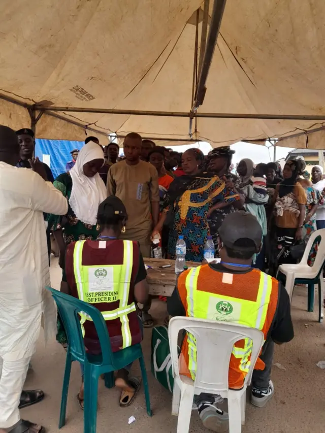 Inec official siddon for chair while voters dey stand for line.