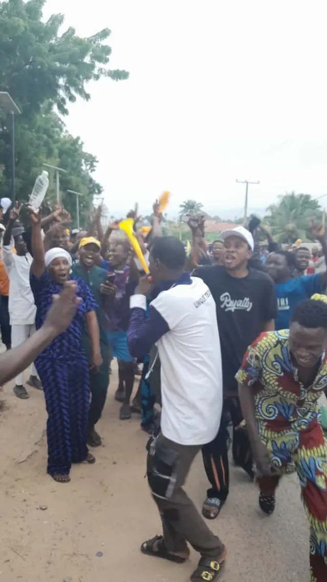 Crowd of pipo dey dance wit pesin wey dey blow yellow plastic trumpet