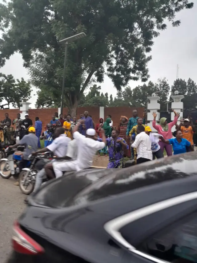 Pipo wey wear white dey ontop okada dey happy as dem drive dey pass pipo wey dey ground wey dey happy too
